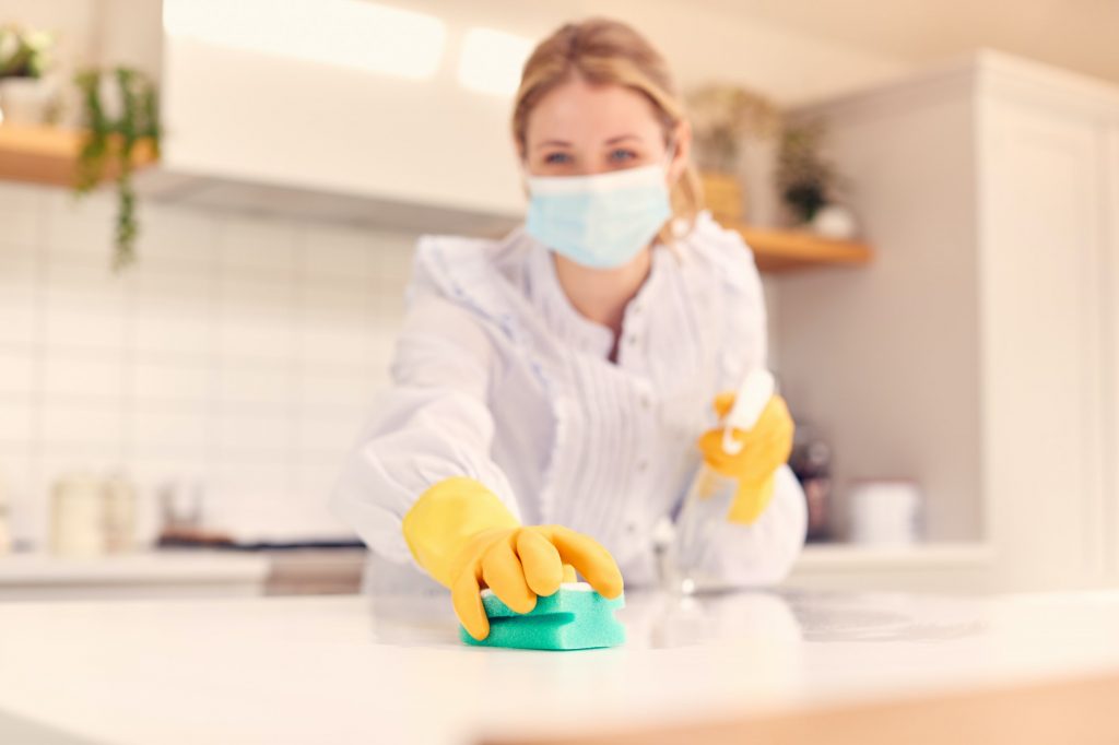 Woman At Home Wearing Mask In Kitchen Doing Housework And Cleaning Counter Surface With Spray